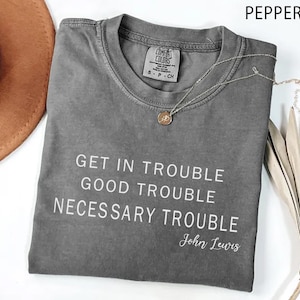 May include: A gray Comfort Colors t-shirt with the quote "Get in Trouble, Good Trouble, Necessary Trouble" by John Lewis. A silver necklace with a pendant and a brown hat are also visible.