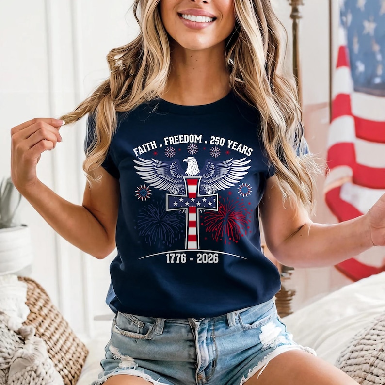 May include: Navy blue t-shirt with a patriotic design. The shirt features a cross with the American flag, an eagle, and the text "Faith, Freedom, 250 Years" and "1776-2026". Red and blue fireworks are also included in the design.