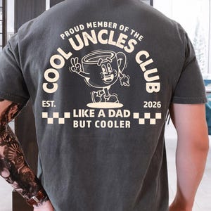 May include: Dark gray t-shirt with white text and graphic. The shirt reads "COOL UNCLES CLUB" with a cartoon coffee cup character. Below the graphic, it says "LIKE A DAD BUT COOLER" and "EST. 2026".