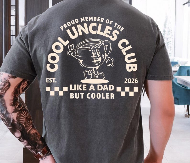 May include: Dark gray t-shirt with white text and graphic. The shirt reads "COOL UNCLES CLUB" with a cartoon coffee cup character. Below the graphic, it says "LIKE A DAD BUT COOLER" and "EST. 2026".