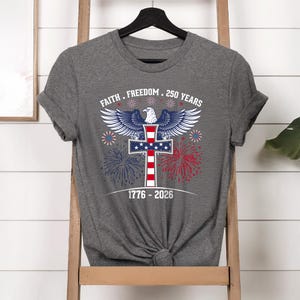 May include: Gray t-shirt featuring a patriotic design with an eagle, cross, and fireworks. The text reads "Faith. Freedom. 250 Years" and "1776-2026". The design incorporates red, white, and blue colors.