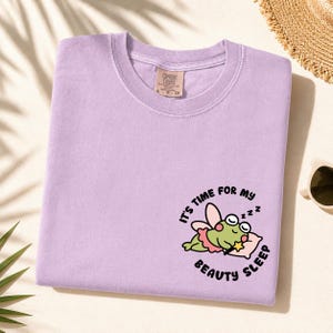 May include: A light purple crewneck t-shirt featuring a graphic of a green frog with pink fairy wings and a skirt sleeping on a pillow. The frog holds a yellow star wand, and the text reads, "IT'S TIME FOR MY BEAUTY SLEEP" in black lettering.
