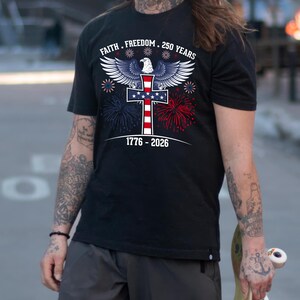 May include: Black t-shirt with a patriotic design featuring an eagle, cross, and fireworks in red, white, and blue. Text includes "Faith. Freedom. 250 Years" and "1776-2026".