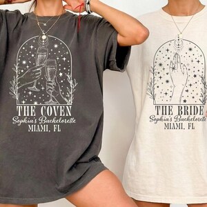 May include: A charcoal gray t-shirt with a white graphic of hands holding champagne flutes under an arch with stars and the text THE COVEN Sophia's Bachelorette MIAMI, FL, and a cream t-shirt with a hand graphic and THE BRIDE Sophia's Bachelorette MIAMI, FL.