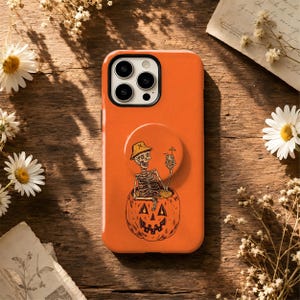 May include: An orange phone case featuring a black line art illustration of a skeleton wearing a bucket hat and holding a drink while sitting inside a carved jack-o'-lantern pumpkin. The case has a circular grip mount attached to the back.