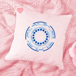 Pu&ograve; includere: Un cuscino quadrato rosa chiaro con un design tecnologico circolare blu e azzurro. Il design presenta cerchi concentrici con un motivo quadrato centrale e linee di collegamento. Un piccolo cuore rosa &egrave; attaccato.