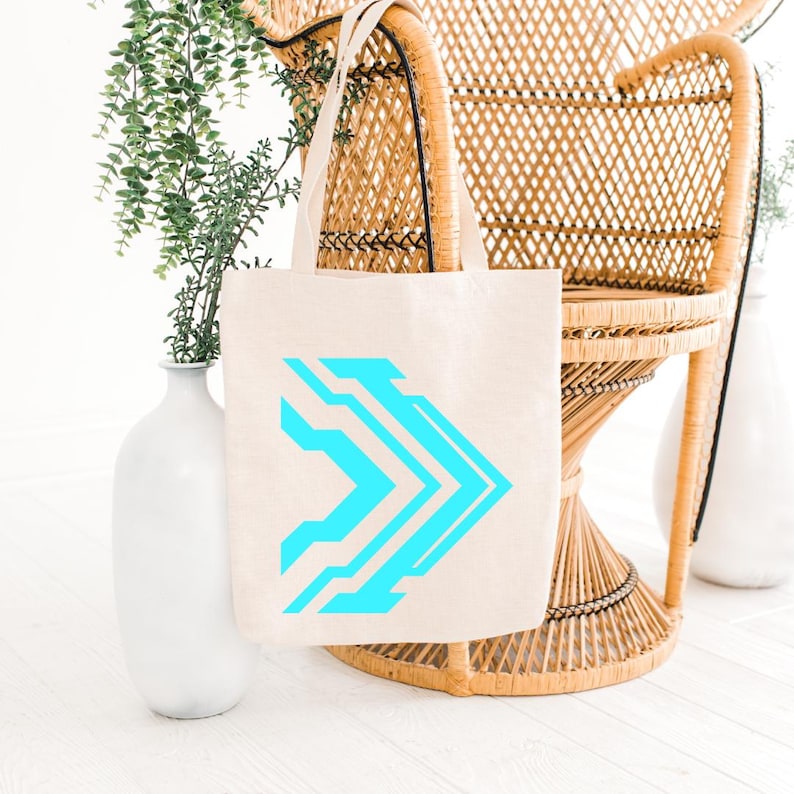 Pu&ograve; includere: Una borsa tote in tela beige con un design a freccia geometrica blu brillante. La borsa ha uno stile semplice e minimalista con manici robusti. La borsa &egrave; esposta vicino a una sedia in vimini e vasi bianchi con vegetazione.