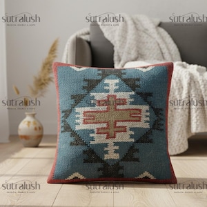 May include: Square throw pillow with a geometric pattern in shades of blue, red, and beige. The pillow has a textured appearance and a red border. The pillow is placed on a couch.