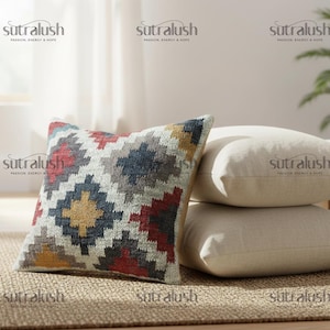 May include: A decorative throw pillow with a geometric pattern in red, blue, yellow, and gray. The pillow is placed on a woven rug next to a stack of solid cream-colored pillows. The brand name "sutralush" is visible.