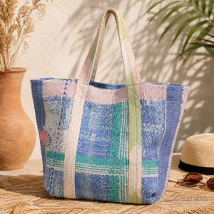 May include: A quilted tote bag featuring a patchwork design in shades of blue, teal, and soft pink. The bag has two sturdy, light-colored fabric handles and a textured surface with visible stitching patterns throughout the fabric construction.