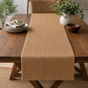 May include: A rectangular table runner featuring a solid tan color and a dense, uniform knit texture. The runner is positioned lengthwise down the center of a wooden table, showcasing a consistent vertical ribbed pattern throughout its length.