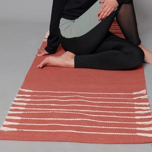 May include: A woman is seated on a terracotta-colored yoga mat with white stripes. She wears a black hoodie with the Nike logo and black and gray leggings. The mat is rectangular and has a fringed edge.