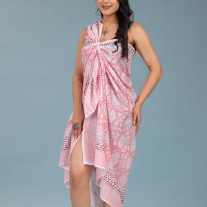 May include: A light pink and white beach wrap featuring a floral and striped pattern, styled as a multiway garment. The fabric is draped and knotted at the shoulder, creating a sleeveless silhouette with a side slit. Text reads Multiway Beach Wrap Styling.