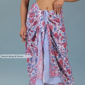 May include: A white beach wrap and pareo featuring a repeating floral pattern in shades of pink and blue, accented by vertical light blue and white striped borders along the edges. The fabric is draped to form a skirt with a central knotted detail.