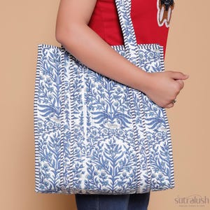 May include: A blue and white floral print tote bag with comfortable shoulder straps. The bag features a quilted design with a repeating pattern of stylized flowers and leaves. The straps are white with a blue stripe. The bag is held by a person wearing a red shirt.