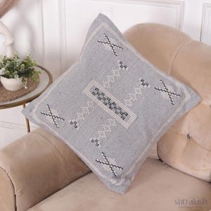 May include: A square, light gray decorative pillow with a geometric pattern in black and white. The pillow is placed on a beige sofa, showcasing its design and texture. The pillow's dimensions are not visible.