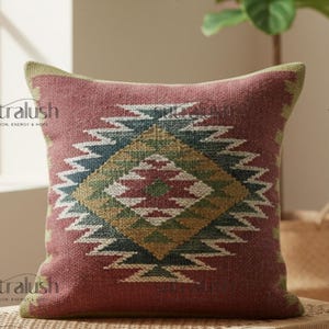 May include: A square decorative pillow with a geometric pattern. The pillow has a reddish-brown background with a central diamond design in shades of green, blue, and white. The edges are trimmed with a green border. The pillow is placed on a wooden surface.