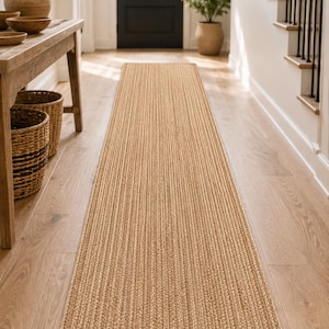 May include: A long, rectangular runner rug featuring a natural tan color and a braided texture. The rug is constructed from woven fibers, creating a consistent, repeating pattern of parallel braids that run the entire length of the floor covering.