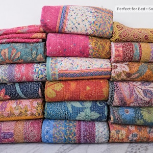 May include: A stack of folded kantha quilts featuring various colorful patterns, including floral, geometric, and abstract designs in shades of pink, blue, orange, yellow, and green. The top quilt has a label reading Perfect for Bed • Sofa • Gift.