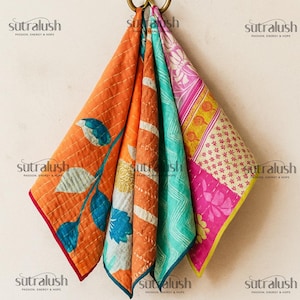 May include: Four colorful, patterned fabric squares hang from a brass ring. The squares feature orange, teal, and pink hues with floral and geometric designs. The edges are trimmed in contrasting colors, and the fabric is quilted.