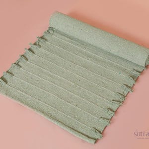 Puede incluir: Una alfombra tejida verde salvia enrollada con un borde con flecos. La alfombra tiene una textura acanalada y se muestra sobre una superficie rosa. La alfombra es un artículo de decoración del hogar.