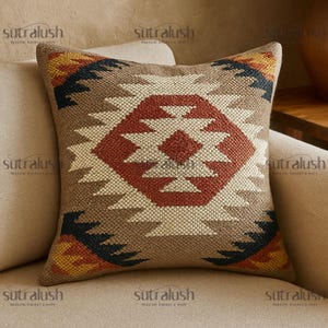 May include: Square decorative pillow with a geometric pattern in shades of brown, red, cream, black, and yellow. The pillow features a central diamond design and is placed on a beige sofa. The pillow has the text "sutralush" on it.