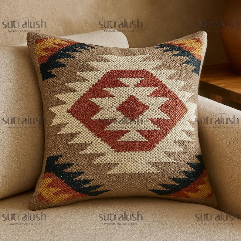 May include: Square decorative pillow with a geometric pattern in shades of brown, red, cream, black, and yellow. The pillow features a central diamond design and is placed on a beige sofa. The pillow has the text "sutralush" on it.