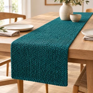 May include: A long, rectangular teal table runner featuring a thick, braided knit texture. The runner is centered on a light-toned wooden dining table, accompanied by white ceramic plates, a textured vase, and a small bowl.