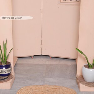 May include: Oval-shaped, natural jute rug. The rug is light brown and sits on a gray tiled floor. Two potted plants flank the rug, and a light pink wall is in the background. The text "Reversible Design" is visible.
