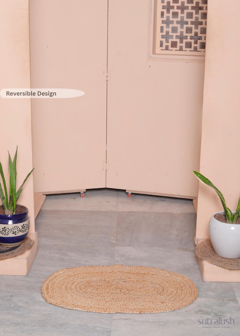 May include: Oval-shaped, natural jute rug. The rug is light brown and sits on a gray tiled floor. Two potted plants flank the rug, and a light pink wall is in the background. The text "Reversible Design" is visible.