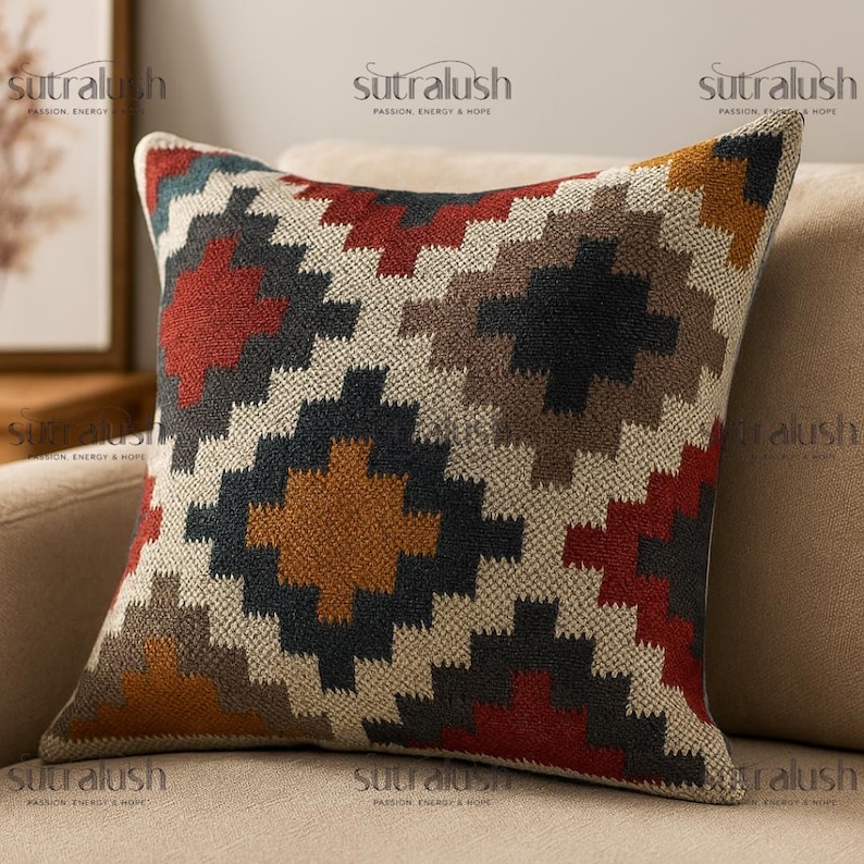 May include: Square throw pillow with a geometric pattern in shades of red, black, brown, and gold on a cream background. The pillow is made of a textured fabric and has a neutral-colored border.