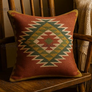 May include: Square decorative pillow with a geometric pattern in shades of red, green, yellow, and cream. The pillow is placed on a wooden chair, showcasing its textured design and warm color palette, suitable for home decor.