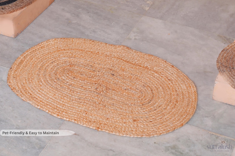 May include: Oval-shaped, natural-toned jute rug. The rug is made of tightly woven fibers, creating a textured surface. The rug is placed on a light gray tiled floor. The text "Pet-Friendly & Easy to Maintain" is visible.