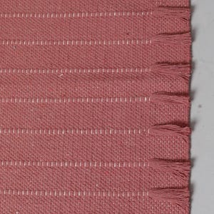 Op de afbeelding: Een geweven kleed in een oudroze kleur met een textuurrijk stoffen oppervlak, horizontale witte stiklijnen en een reeks bijpassende roze franjes langs de rechterrand. De merknaam sutralush staat in de hoek van het kleed gedrukt.