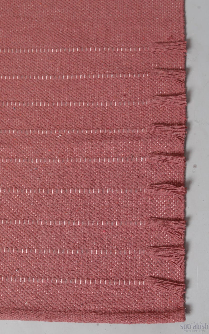 Op de afbeelding: Een geweven kleed in een oudroze kleur met een textuurrijk stoffen oppervlak, horizontale witte stiklijnen en een reeks bijpassende roze franjes langs de rechterrand. De merknaam sutralush staat in de hoek van het kleed gedrukt.