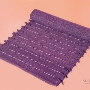 May include: A rolled-up purple woven mat with white stripes and fringed edges. The mat is made of a textured fabric and is displayed on a peach-colored surface. The mat is a home decor item.