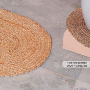 May include: Two hand-braided jute mats, one oval and one round, sit on a marble surface. The oval mat is in the foreground, while the round mat is partially obscured. Text reads "Hand-Braided from 100% Natural Jute."