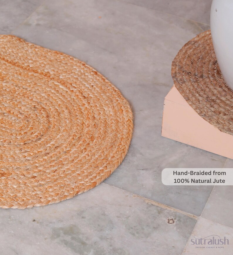 May include: Two hand-braided jute mats, one oval and one round, sit on a marble surface. The oval mat is in the foreground, while the round mat is partially obscured. Text reads "Hand-Braided from 100% Natural Jute."