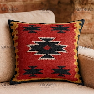 May include: Square decorative pillow with a geometric pattern. The pillow features a red background with black and white geometric shapes. The border is black and yellow. The pillow is placed on a beige sofa.
