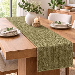 May include: A long, rectangular olive green table runner with a thick, braided, woven texture. The runner is centered on a wooden dining table, featuring a white vase with greenery, a candle in a woven holder, and place settings.