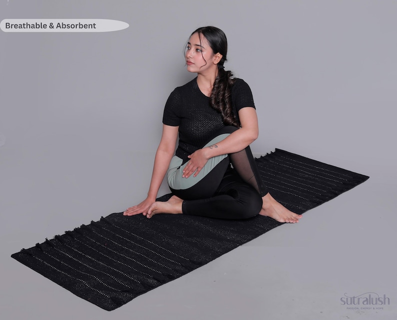 Op de afbeelding: Een zwarte yogamat met een getextureerd oppervlak en franjes aan de randen. De mat bevat de tekst "Breathable & Absorbent" in de linkerbovenhoek en het "sutralush"-logo in de rechterbenedenhoek, op een neutrale grijze achtergrond.