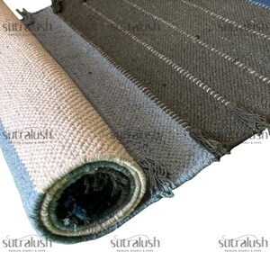 Peut inclure: Tapis enroul&eacute;, multicolore, avec un tissage textur&eacute;. Le tapis pr&eacute;sente des nuances de gris, de bleu et de beige, avec des coutures blanches et des franges. Le nom de la marque "Sutralush" est imprim&eacute;.