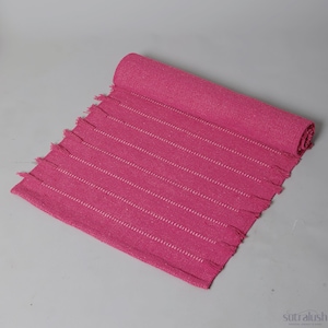 May include: A rolled-up, hot pink yoga mat with white stripes and fringe detailing. The mat is made of woven material and is partially unrolled, displaying the striped pattern. The mat is designed for yoga and fitness activities.