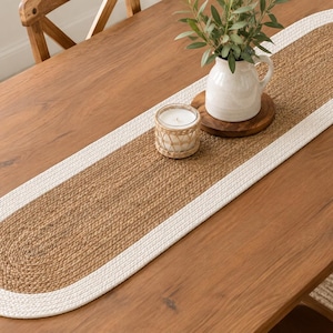 May include: An oval table runner featuring a textured brown jute center bordered by a thick white braided rope edge. The runner is placed on a wooden table and holds a white ceramic pitcher with green foliage and a small candle in a woven holder.