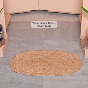May include: An oval-shaped, woven jute rug in natural tan tones. The rug is centered on a gray tiled floor, with potted plants on either side. Text in the image reads: "Warm, Natural Texture for Any Space."