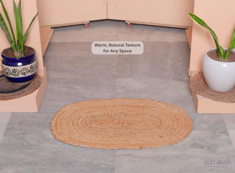 May include: An oval-shaped, woven jute rug in natural tan tones. The rug is centered on a gray tiled floor, with potted plants on either side. Text in the image reads: "Warm, Natural Texture for Any Space."