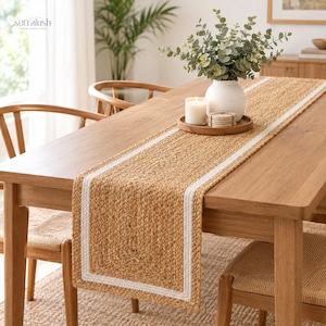 May include: A rectangular table runner made of braided natural tan jute fibers with a thick white fabric border. The runner is centered on a wooden dining table, featuring a woven texture and a clean, structured rectangular shape.