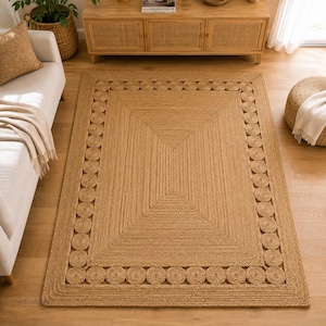 May include: A rectangular area rug crafted from natural tan jute fibers. The rug features a central rectangular section surrounded by a border of repeating circular jute coils, all framed by a solid, braided outer edge in a matching tan tone.