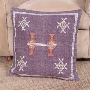 May include: Square purple decorative pillow with a geometric design. The pillow features a central motif of two orange vases connected by a white line, surrounded by white and gold starburst patterns. The border is white.