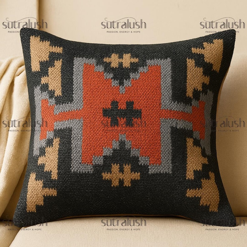 May include: Square decorative pillow with a geometric pattern. The pillow features a black background with a central design in shades of orange, gray, and tan. The pillow is placed on a beige surface. The text "sutralush" is visible.
